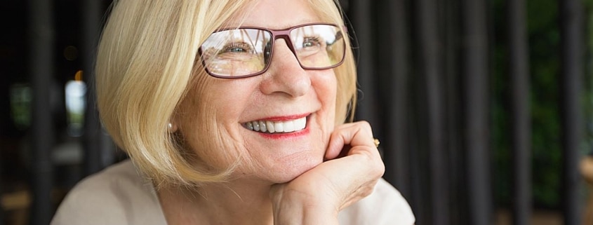 mulher de 60 anos usando óculos sorri na janela com a mão no queixo.